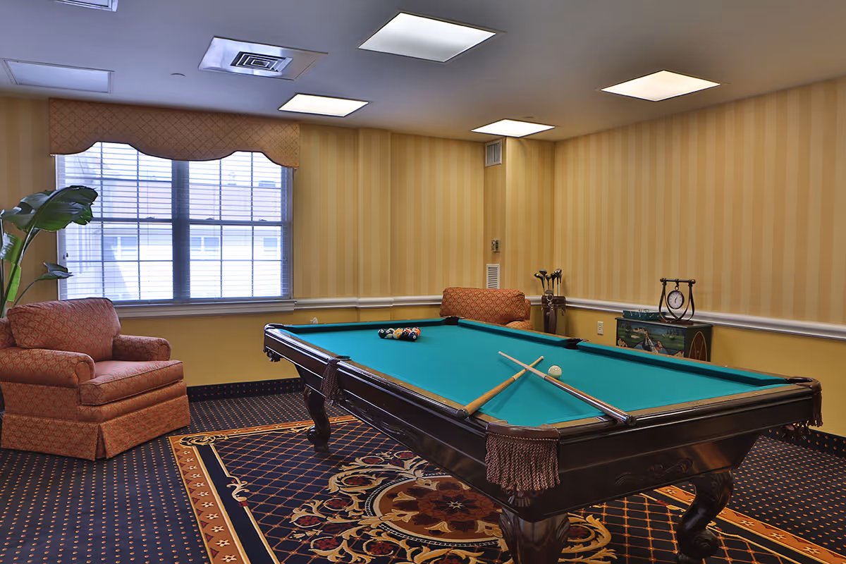 A recreational room with a pool table in the center, two pool cues resting on the table, a rack of pool balls arranged for a game, two upholstered armchairs with patterned fabric, a large window with blinds and a valance, a decorative carpet under the pool table, and a small cabinet with a clock and golf clubs in the corner.