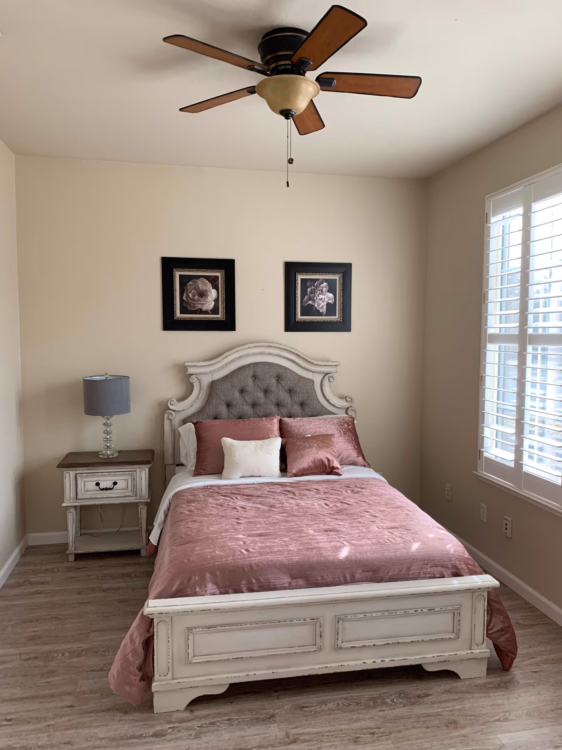 A neatly made bed with a tufted headboard and pink bedding in a bedroom. There is a white nightstand with a lamp on the left side of the bed. Two framed floral pictures hang on the wall above the headboard. A ceiling fan with wooden blades is mounted on the ceiling. A window with white shutters is on the right side of the room, allowing natural light to enter.