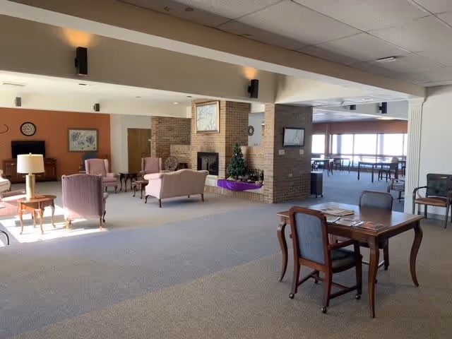 A spacious senior living facility common area with multiple seating arrangements including armchairs and sofas around a brick fireplace. There is a small decorated Christmas tree on the hearth. The room has large windows letting in natural light and a table with chairs in the foreground. The walls are painted white and light brown, and the ceiling has recessed lighting.