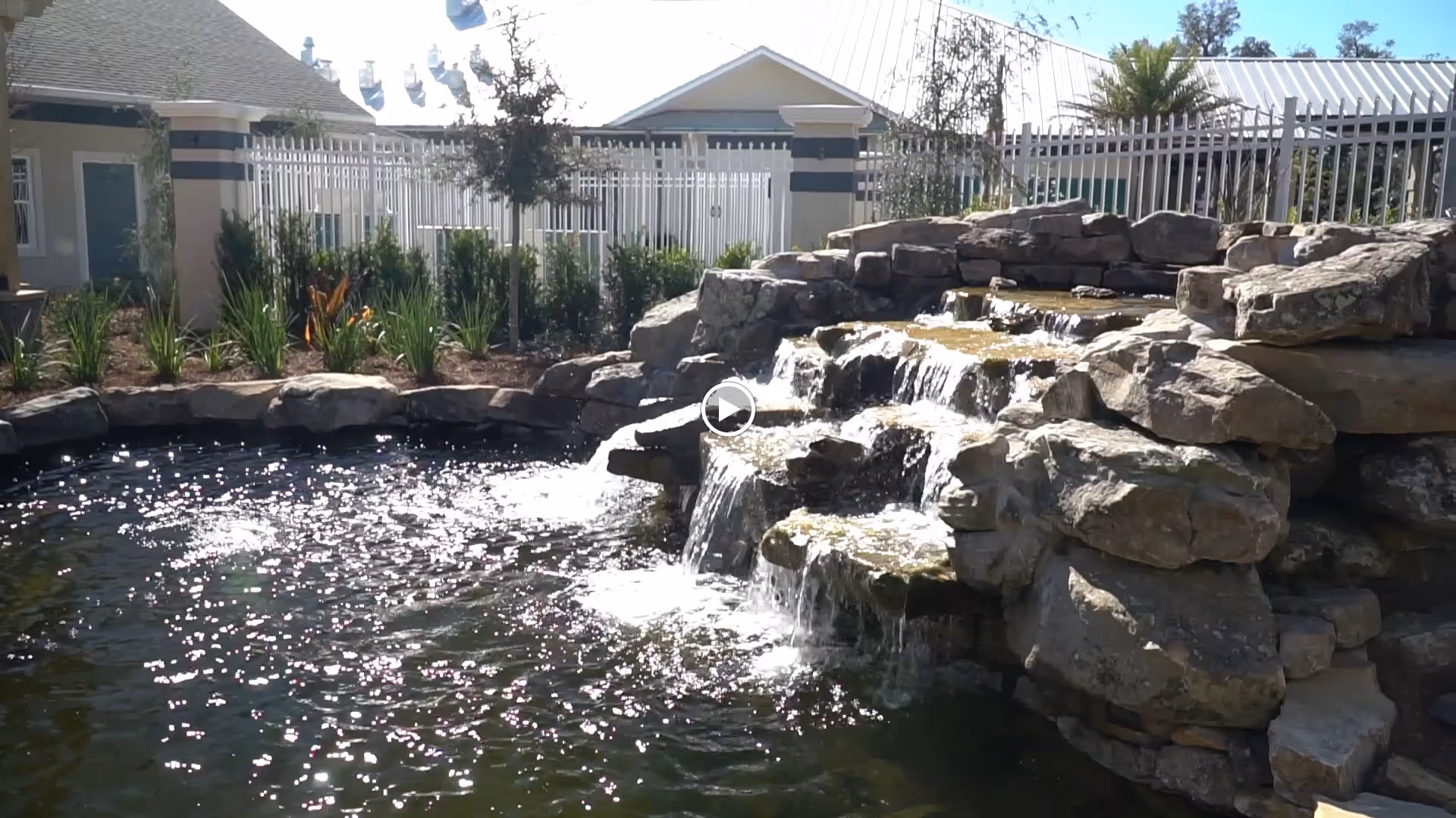 A landscaped outdoor pond with a multi-tiered rock waterfall and buildings with a white fence in the background.