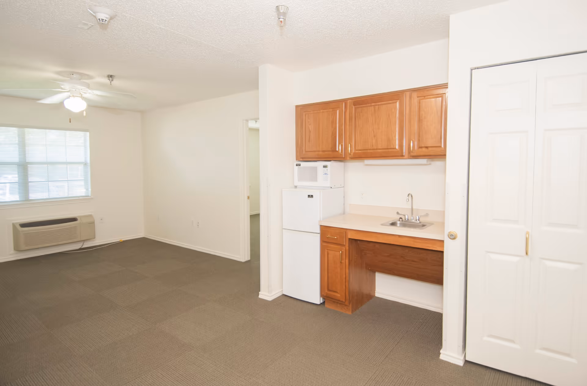 An empty room with beige carpet and white walls featuring a small kitchenette area with wooden cabinets, a white mini refrigerator, a microwave, and a sink. There is a window with blinds, a ceiling fan with light, and a white air conditioning unit mounted below the window. A white double-door closet is visible on the right side.