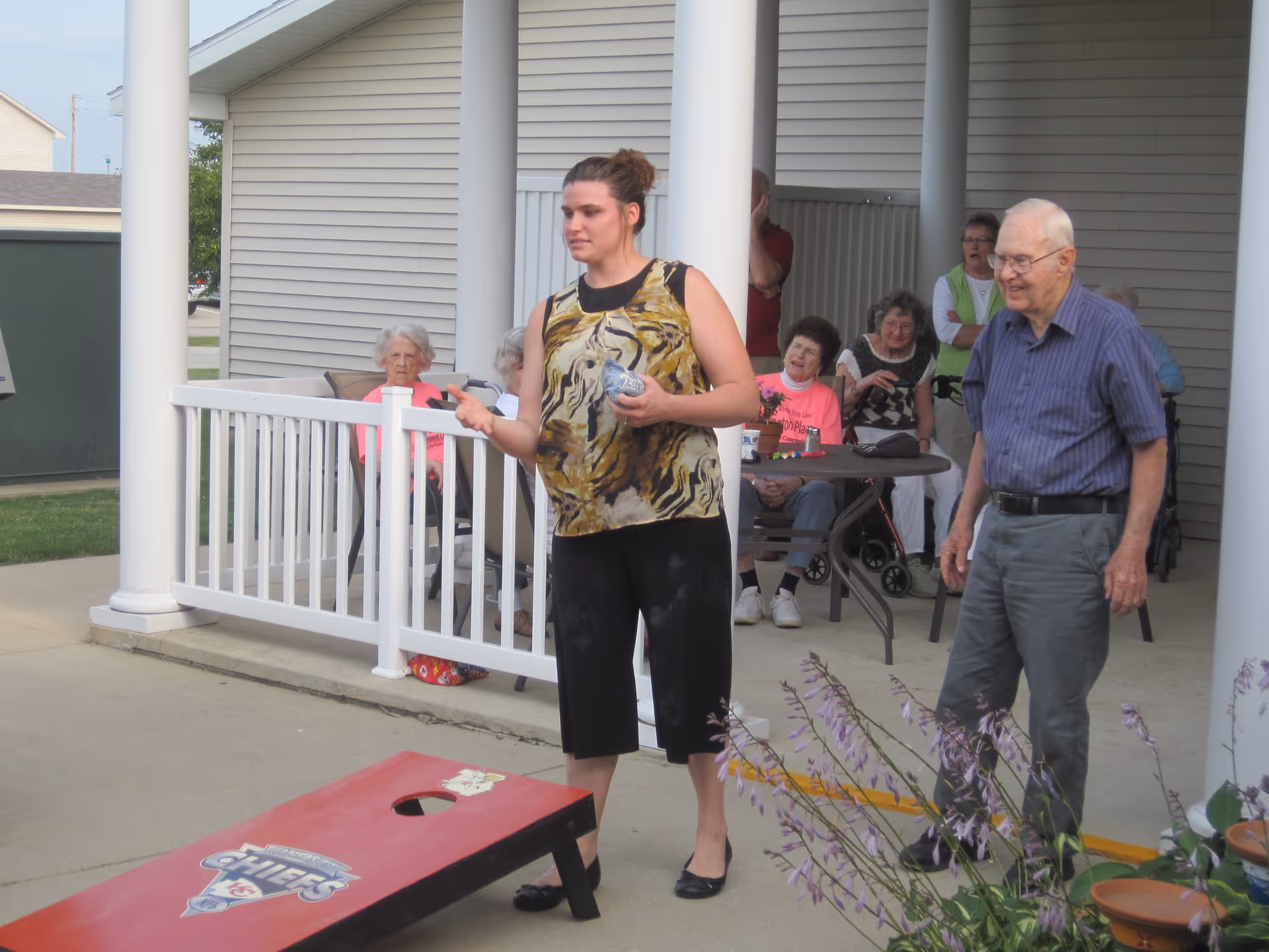 A young woman is playing cornhole outdoors on a concrete patio at Arlington Place at Grundy Center. An elderly man stands nearby watching her. In the background, several elderly people are seated around a table under a covered porch, observing the game. The setting appears to be a senior living facility with a relaxed and social atmosphere.