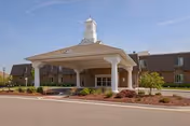 Front entrance of a senior living facility with a covered porte-cochère, white columns, landscaped beds, and a clear blue sky.