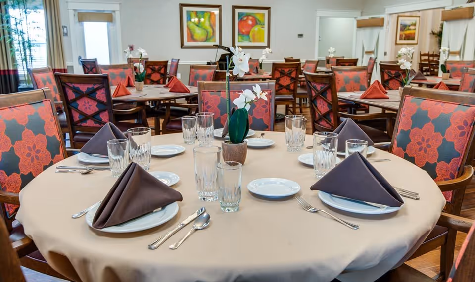 A dining room with multiple round tables set with beige tablecloths, white plates, folded dark napkins, clear drinking glasses, and silverware. Each table has a small potted white orchid as a centerpiece. The chairs have a red and black floral pattern. The room has framed fruit paintings on the walls and large windows with curtains.