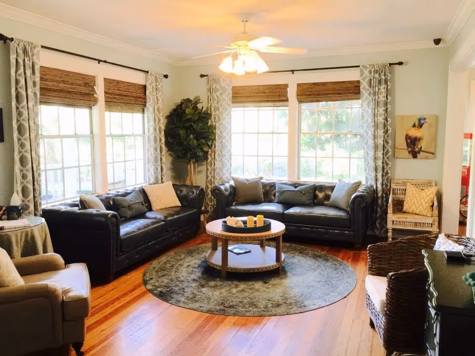 A cozy living room with two black leather sofas, a round wooden coffee table on a circular rug, an armchair, and a wicker chair. The room has large windows with patterned curtains and bamboo blinds, a ceiling fan with lights, a potted plant in the corner, and a painting of a bird on the wall.