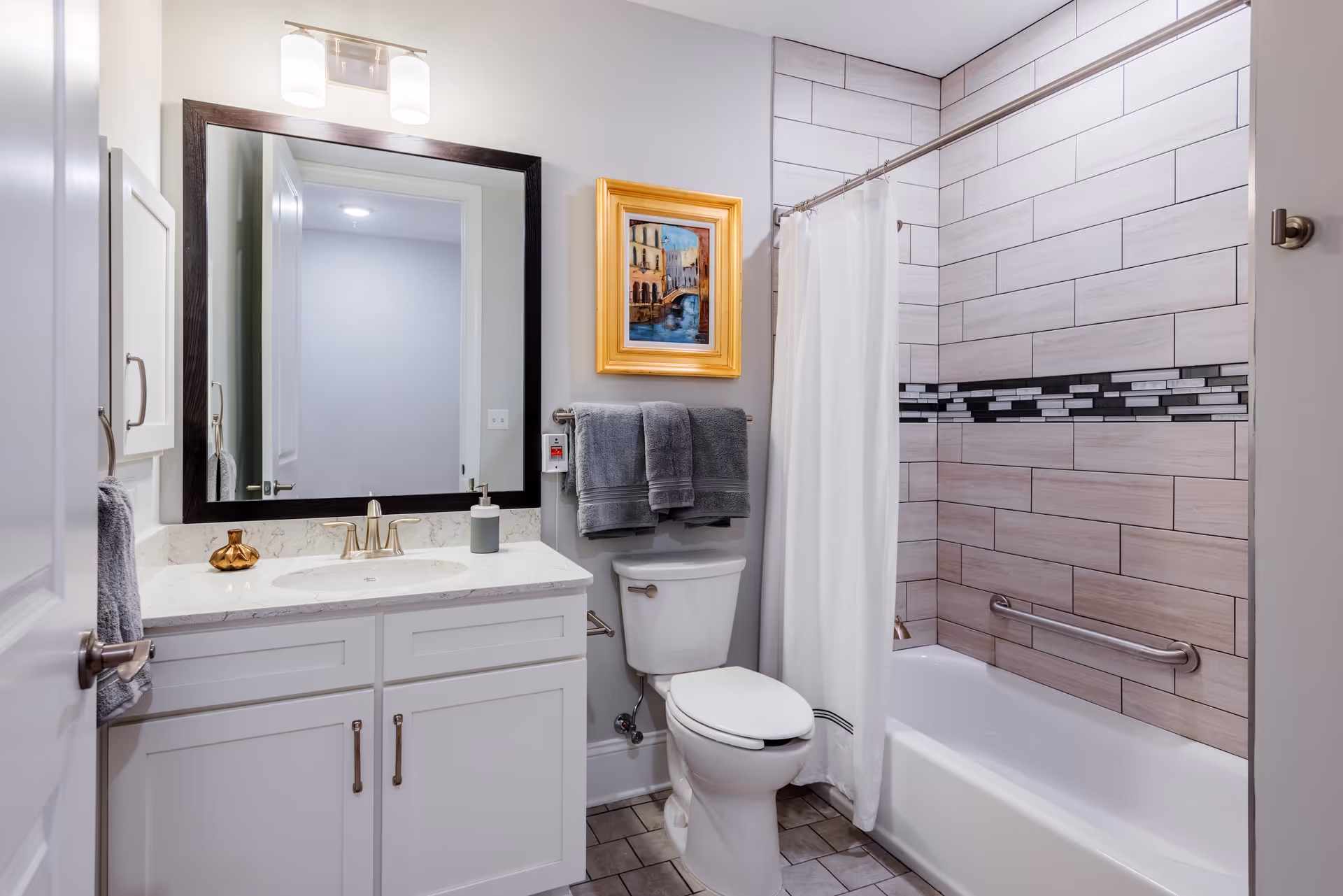 A clean and modern bathroom featuring a white vanity with a marble countertop, a large rectangular mirror with dark frame, a toilet with two gray towels hanging above it, a bathtub with a white shower curtain, and tiled walls with a decorative horizontal strip. There is a small framed painting on the wall above the towels and a silver grab bar inside the bathtub.