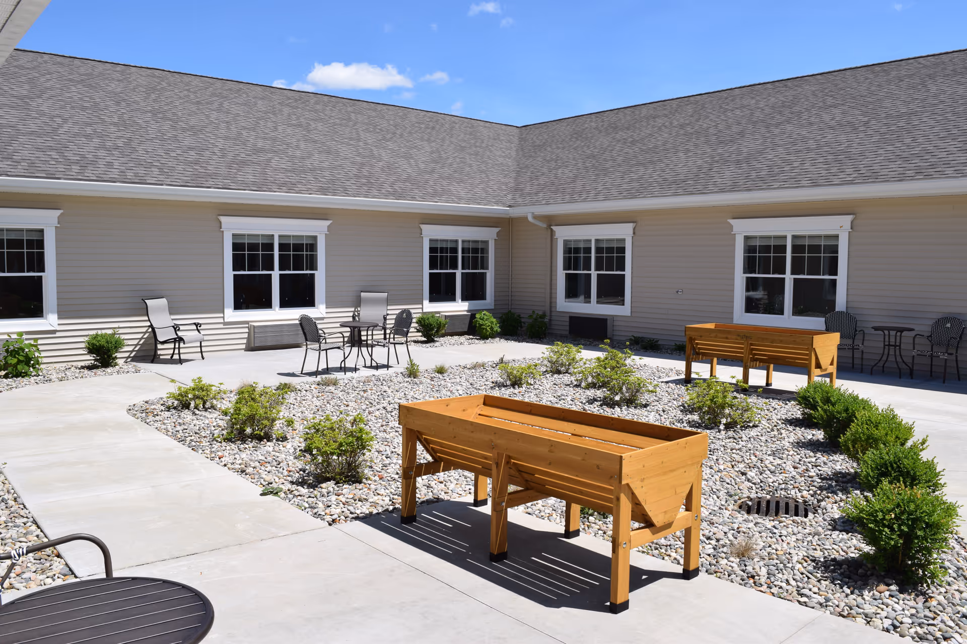 Outdoor courtyard area of Clinton Creek Assisted Living & Memory Care with beige siding building walls, multiple windows, a concrete walkway, small bushes planted in a bed of rocks, two wooden raised garden beds, and several metal chairs and tables arranged around the space under a clear blue sky.