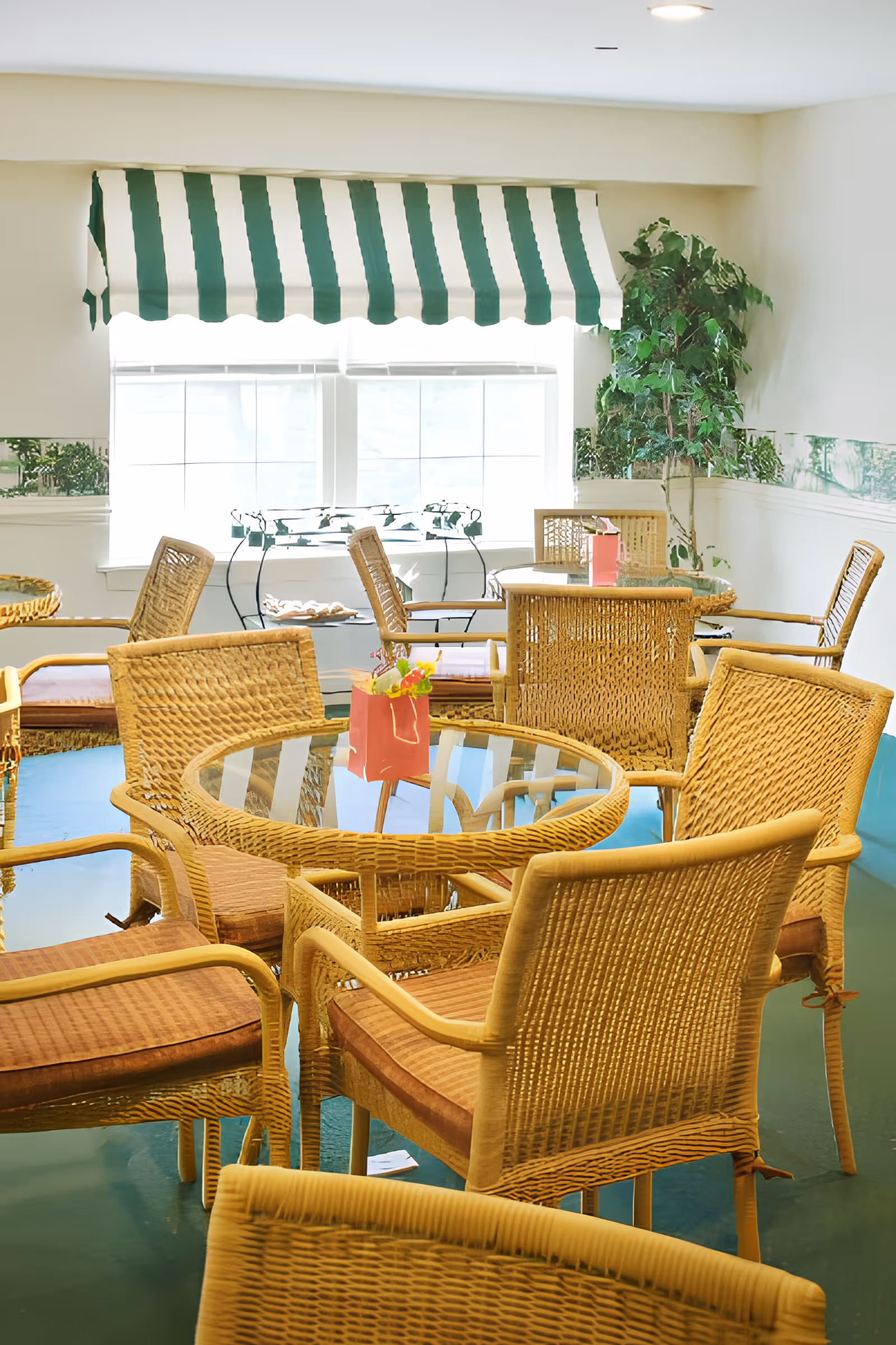 Bright communal seating area with wicker chairs and glass-top tables near a window with a green-and-white striped valance.
