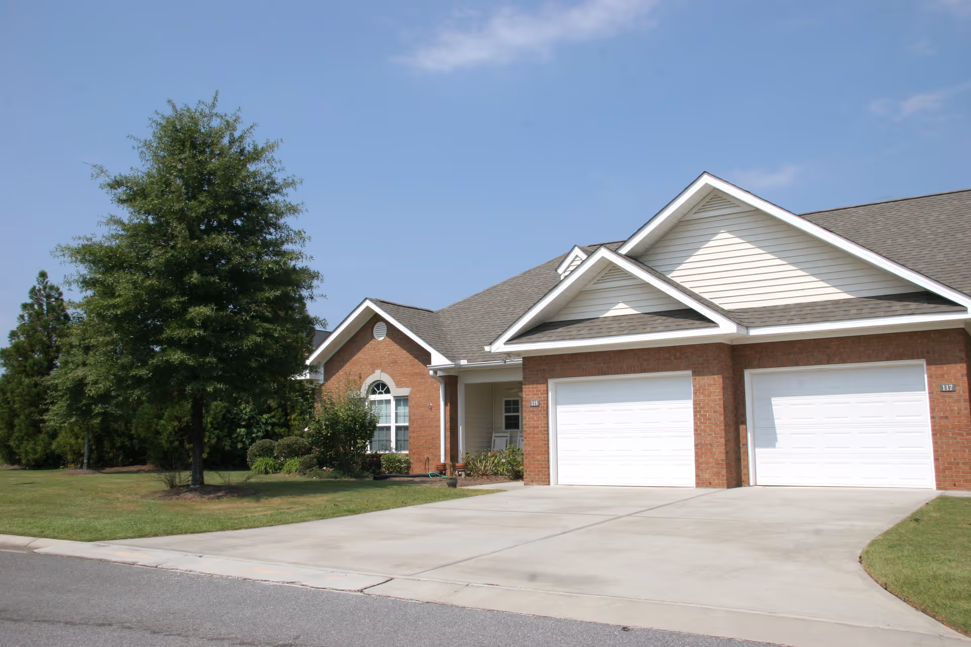 Brick cottage-style duplex with two white garage doors, a front lawn and a tree under a clear blue sky.