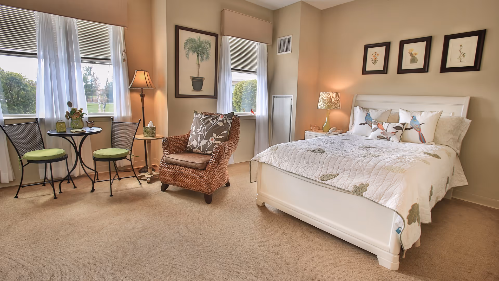 A cozy bedroom with a white bed featuring bird-themed pillows and a quilt. There is a wicker armchair with a floral cushion, a small round table with two chairs with green cushions near two windows with white curtains. The walls are decorated with framed botanical prints and a floor lamp and table lamp provide warm lighting.