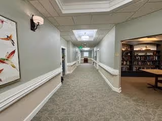 A carpeted hallway in a senior living facility with light green walls, white handrails, and ceiling lights. On the left wall, there is a framed artwork featuring colorful birds. To the right, an open doorway reveals a room with bookshelves and a table.