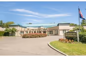 Exterior view of Candlewood Valley Health & Rehabilitation Center showing a single-story building with a green roof, a driveway, landscaped bushes, and an American flag on a flagpole near the entrance.