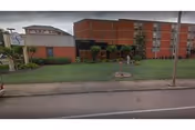 Exterior view of a multi-story brick building with a grassy lawn and a sidewalk in front. The building has several windows and a small covered entrance area. There is a street with a curb and a streetlight visible in the foreground.