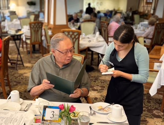 An elderly man sitting at a dining table in a restaurant setting, holding a menu and talking to a waitress who is taking his order. The table is set with white tablecloth, cups, utensils, and a small flower vase. Other diners are visible in the background.