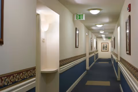 A long indoor hallway with blue carpet and beige walls featuring a floral border. The hallway is lit by ceiling lights and has framed pictures on the walls. There are exit signs visible at the end and along the hallway.