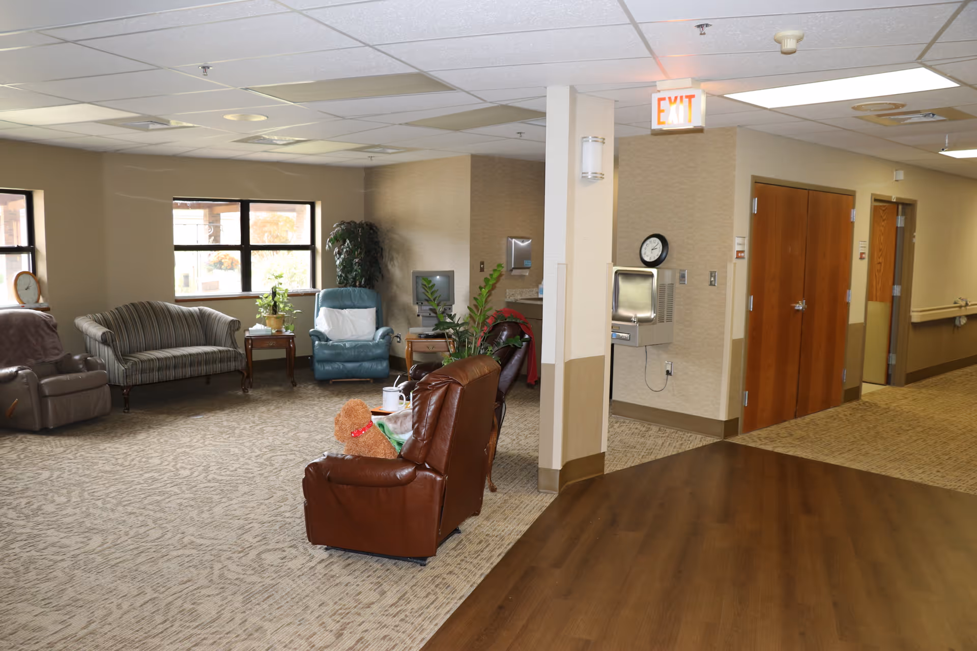 A spacious common area in a senior living facility with several comfortable chairs and a sofa arranged around a small table. There are windows letting in natural light, a water fountain, a clock on the wall, and an exit sign above a hallway entrance. The floor is carpeted with a section of wood flooring near the foreground.