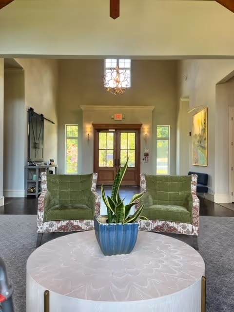 Interior view of a senior living facility lobby with two green upholstered armchairs with patterned sides facing a round white table with a blue pot containing a snake plant. In the background, there is a wooden double door with glass panels, flanked by narrow windows and wall sconces. A chandelier hangs from the ceiling above the door, and there is a painting on the right wall.