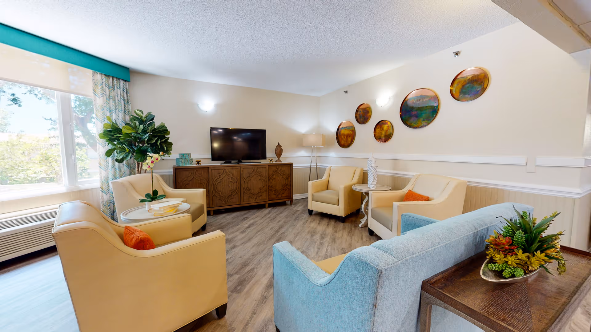 A bright and cozy living room area with a large window letting in natural light. The room features a light blue sofa, three beige armchairs with orange accent pillows, a wooden TV console with a flat-screen TV, and decorative wall art consisting of five circular paintings. There are also side tables with plants and a floor lamp in the corner.