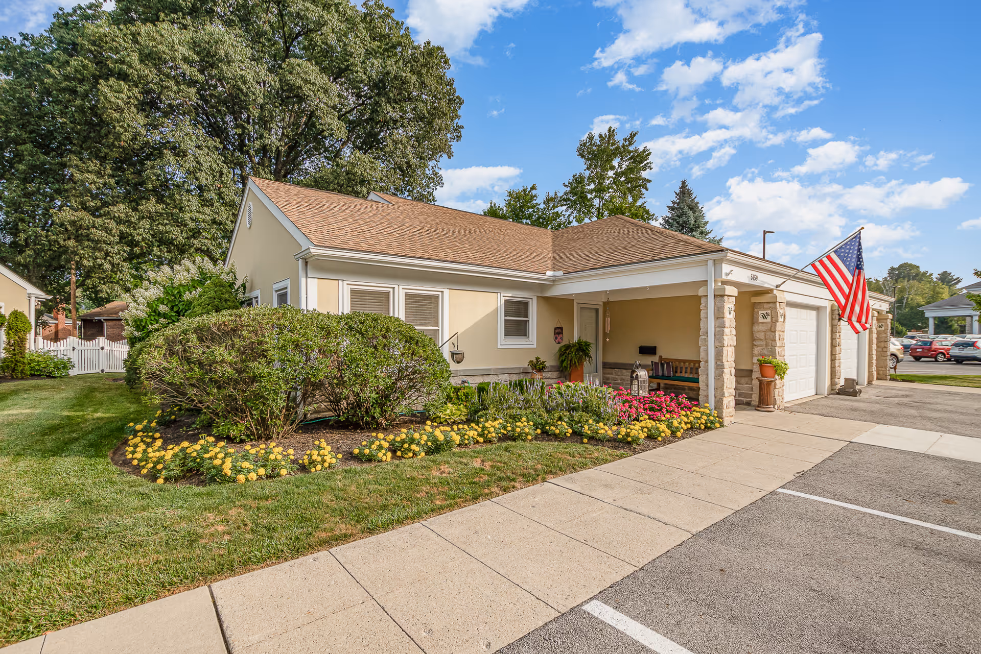 Single-story beige senior living cottage with attached garage, front porch, American flag, and landscaped flowerbeds.