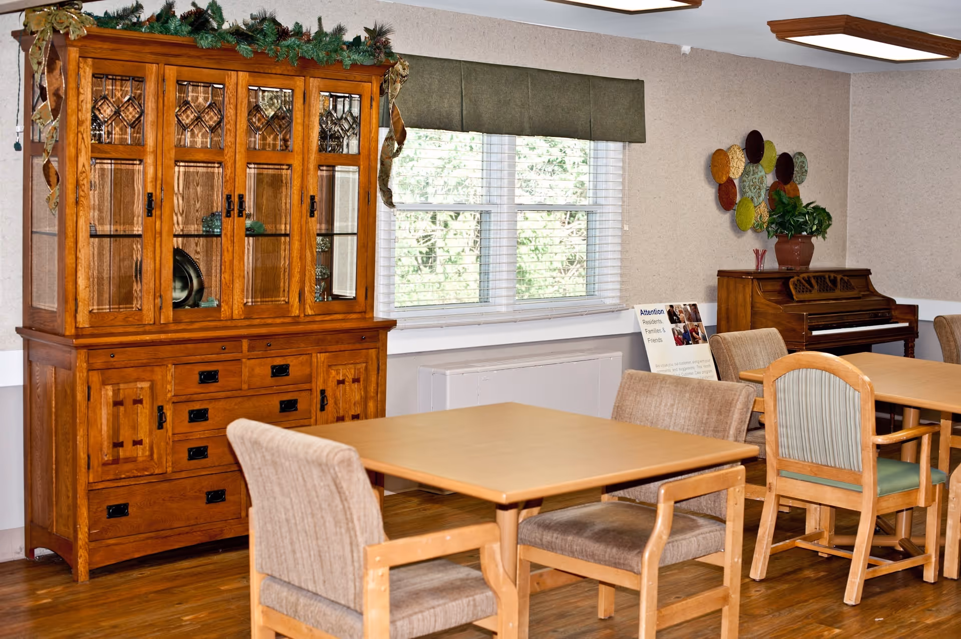 A dining area in a senior living facility featuring wooden tables and cushioned chairs. There is a large wooden cabinet with glass doors on the left side, decorated with greenery on top. A window with white blinds and a green valance is centered on the wall. To the right, there is a wooden piano with a potted plant on top and colorful circular wall decorations above it. The floor is wooden, and the walls are light-colored.