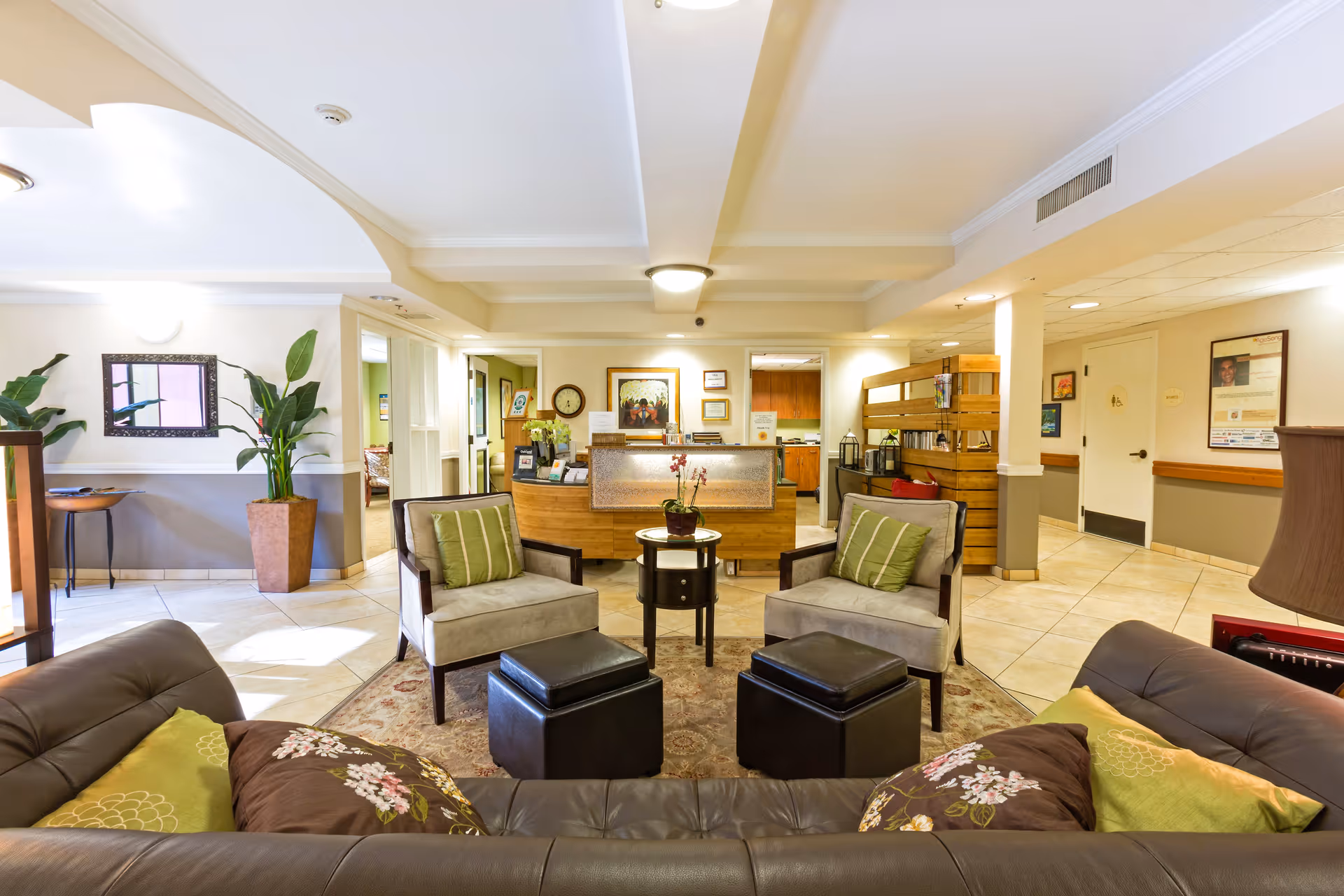 A cozy and well-lit senior living facility lobby area with a brown leather sofa adorned with floral and green pillows in the foreground. Two beige armchairs with green striped pillows face the sofa, separated by a small round table with a potted orchid. Behind the seating area is a wooden reception desk with framed artwork and certificates on the wall. The space features tiled floors, potted plants, and a hallway leading to other rooms.