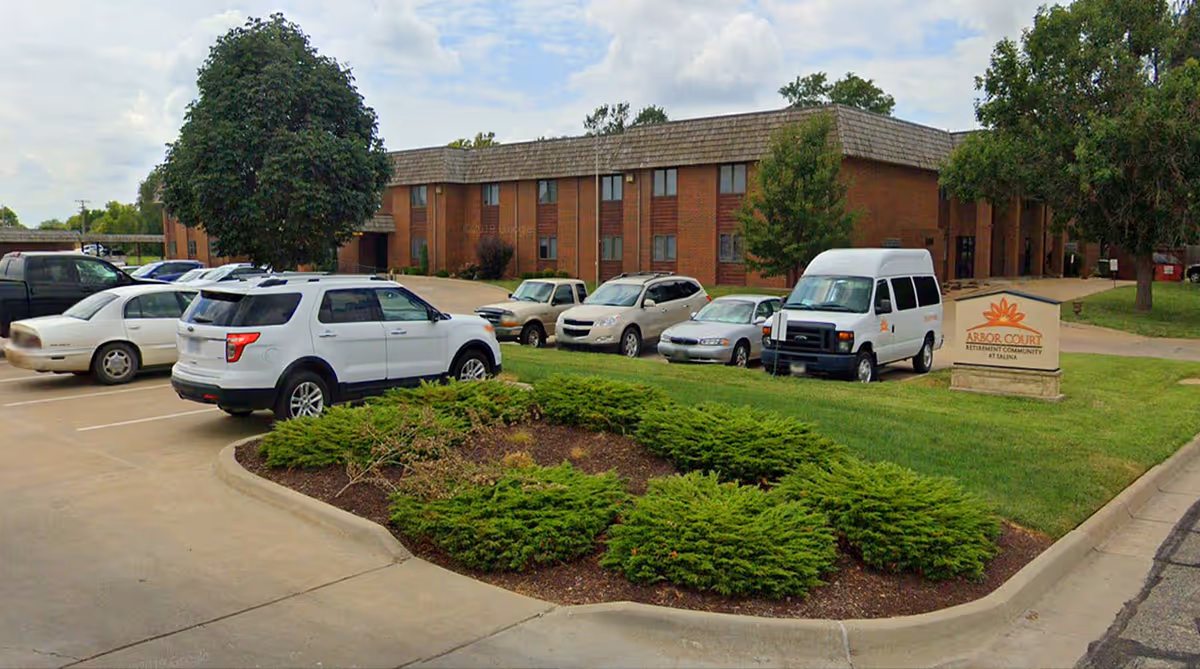 Exterior view of Arbor Court Retirement Community at Salina showing a two-story brick building with multiple windows, a parking lot with several cars and a white van, green landscaped bushes, and a sign with the community's name near the entrance.