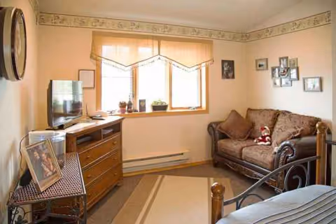 A cozy living room in a senior facility featuring a sofa, TV, dresser, and window.