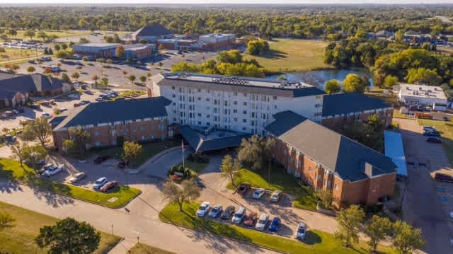 Aerial view of Tealridge Retirement Community showing a large multi-wing building with a mix of brick and white exterior walls, surrounded by parking lots, trees, and green spaces under a clear sky.