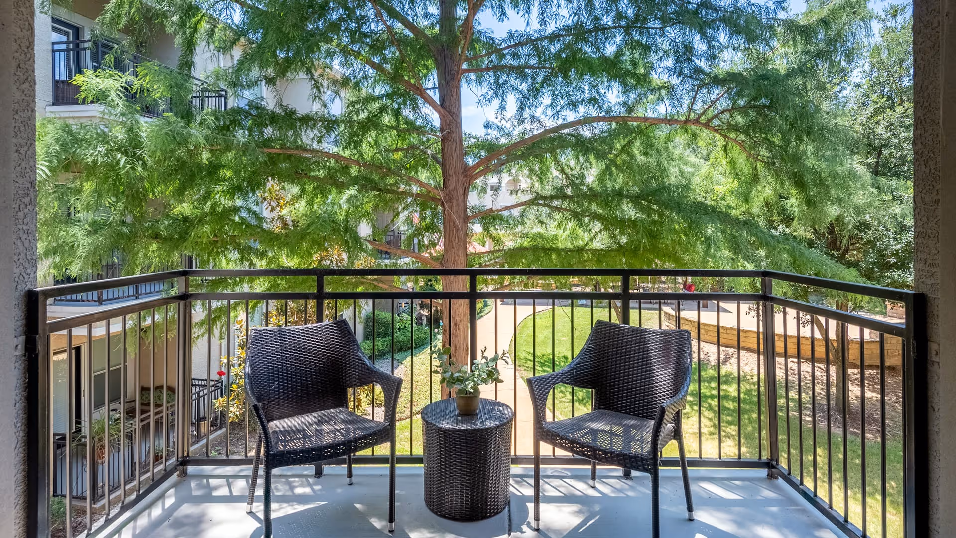 A balcony with two black wicker chairs and a small round table with a potted plant between them, overlooking a green courtyard with trees and a walking path.