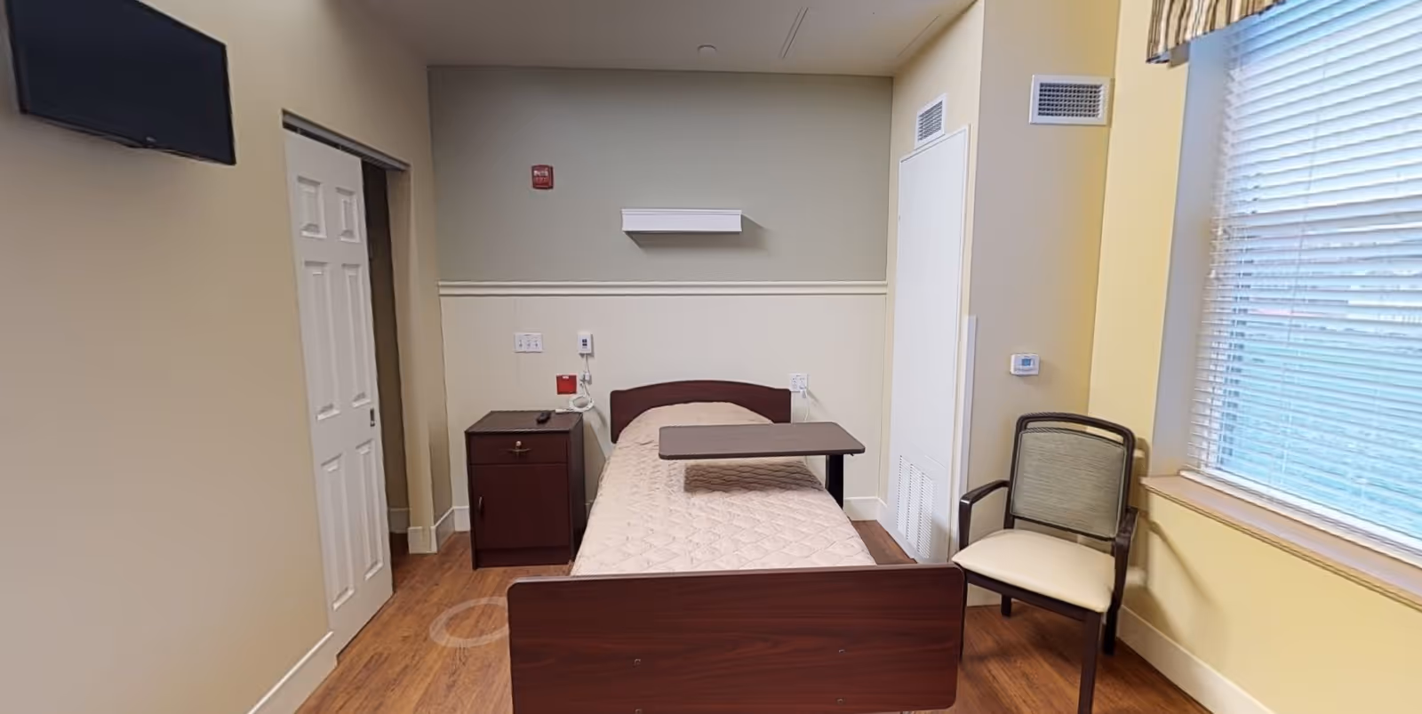 A small bedroom in a senior living facility with a single bed covered with a beige quilted blanket. There is a dark wooden nightstand with a remote control on it next to the bed. A movable overbed table is positioned over the bed. A cushioned chair with wooden arms is placed near a window with blinds. The walls are painted in light colors and the floor has wood-like flooring. A flat-screen TV is mounted on the wall to the left.