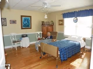 A senior woman sitting in a chair in a well-lit room with a hospital-style bed covered with a blue quilt. The room has wooden floors, a ceiling fan, framed pictures on the walls, a small table with two white chairs, and a window with blue curtains letting in sunlight.