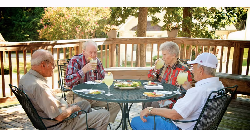 Four elderly residents sit around a round metal patio table on a wooden deck enjoying drinks and salad.