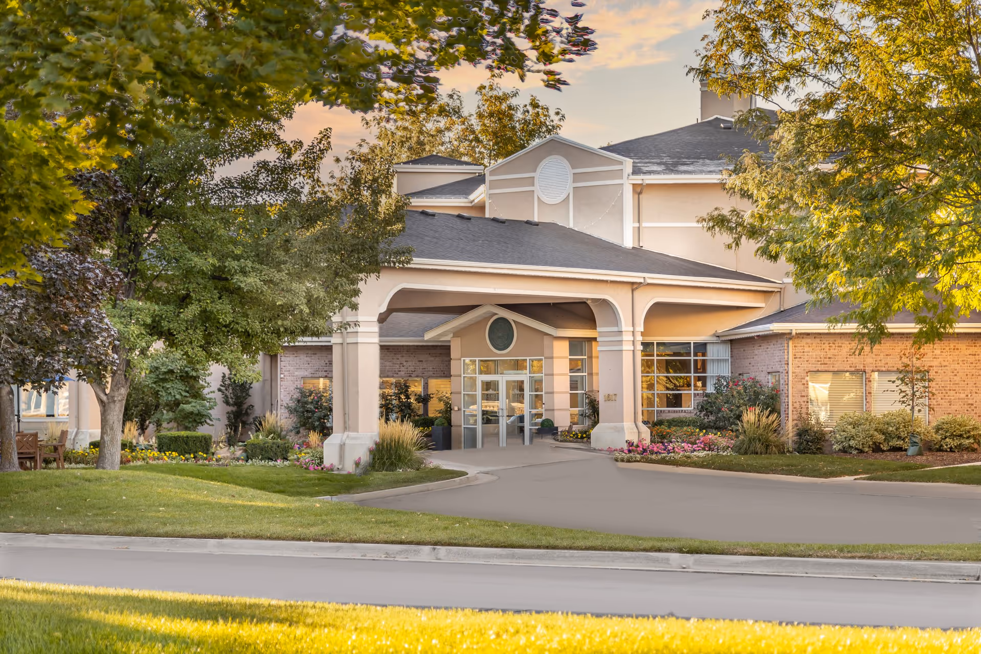 Exterior view of Legacy Retirement Residence building entrance with a covered driveway, surrounded by green trees, bushes, and colorful flowers under a partly cloudy sky at sunset.