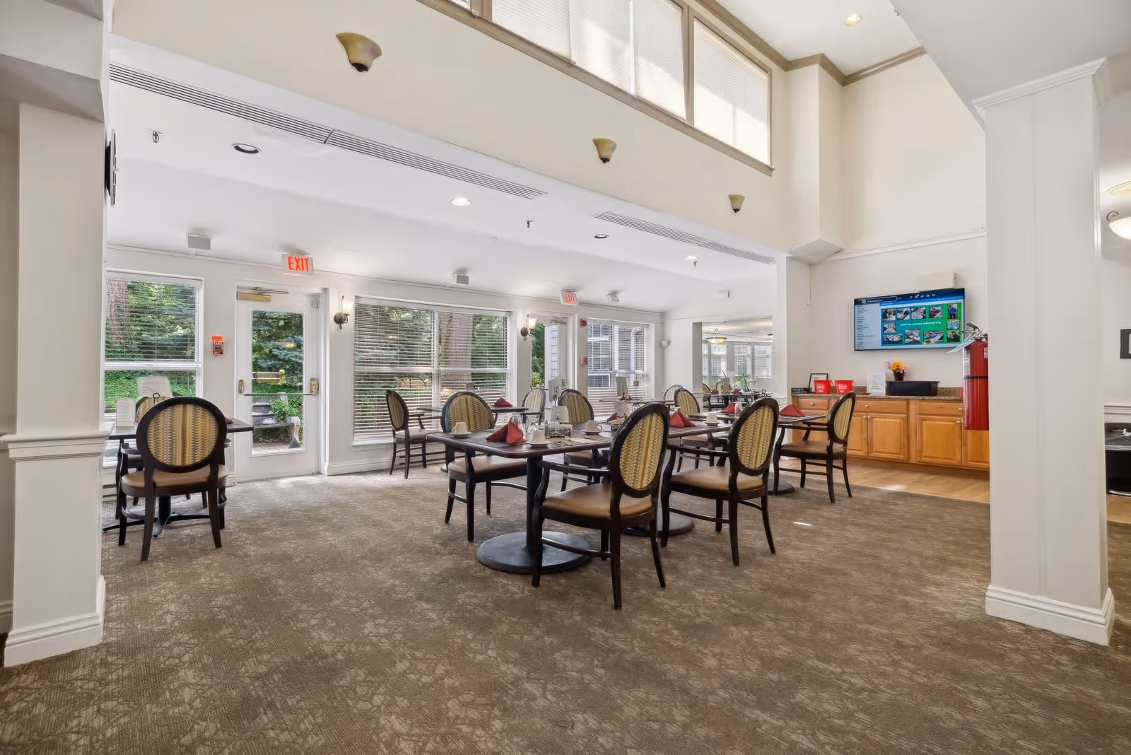 A bright dining room with several tables and chairs arranged neatly. Each table is set with red napkins and glassware. Large windows and glass doors allow natural light to fill the room, and there is a television mounted on the wall above wooden cabinets. The room has a carpeted floor and white walls with high ceilings.