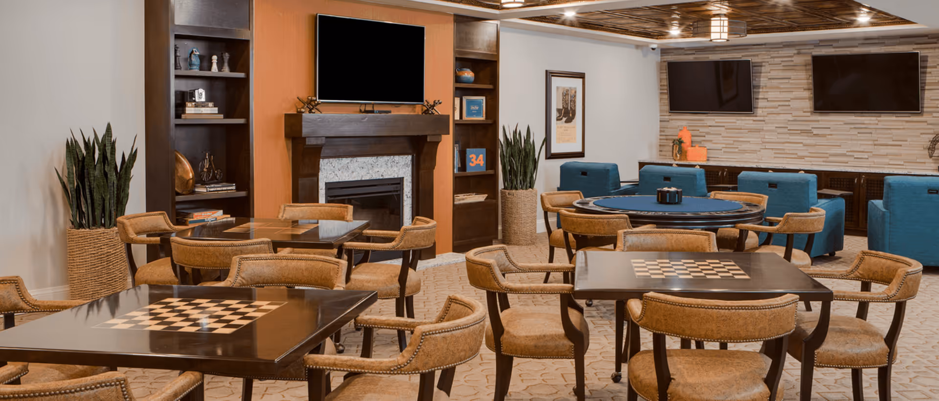 A cozy common area with multiple tables featuring built-in chessboards, surrounded by cushioned chairs. The room includes a fireplace with a mounted TV above it, built-in shelves with decorative items, and additional seating with blue armchairs. The walls are decorated with framed artwork and the ceiling has a wooden design with recessed lighting.