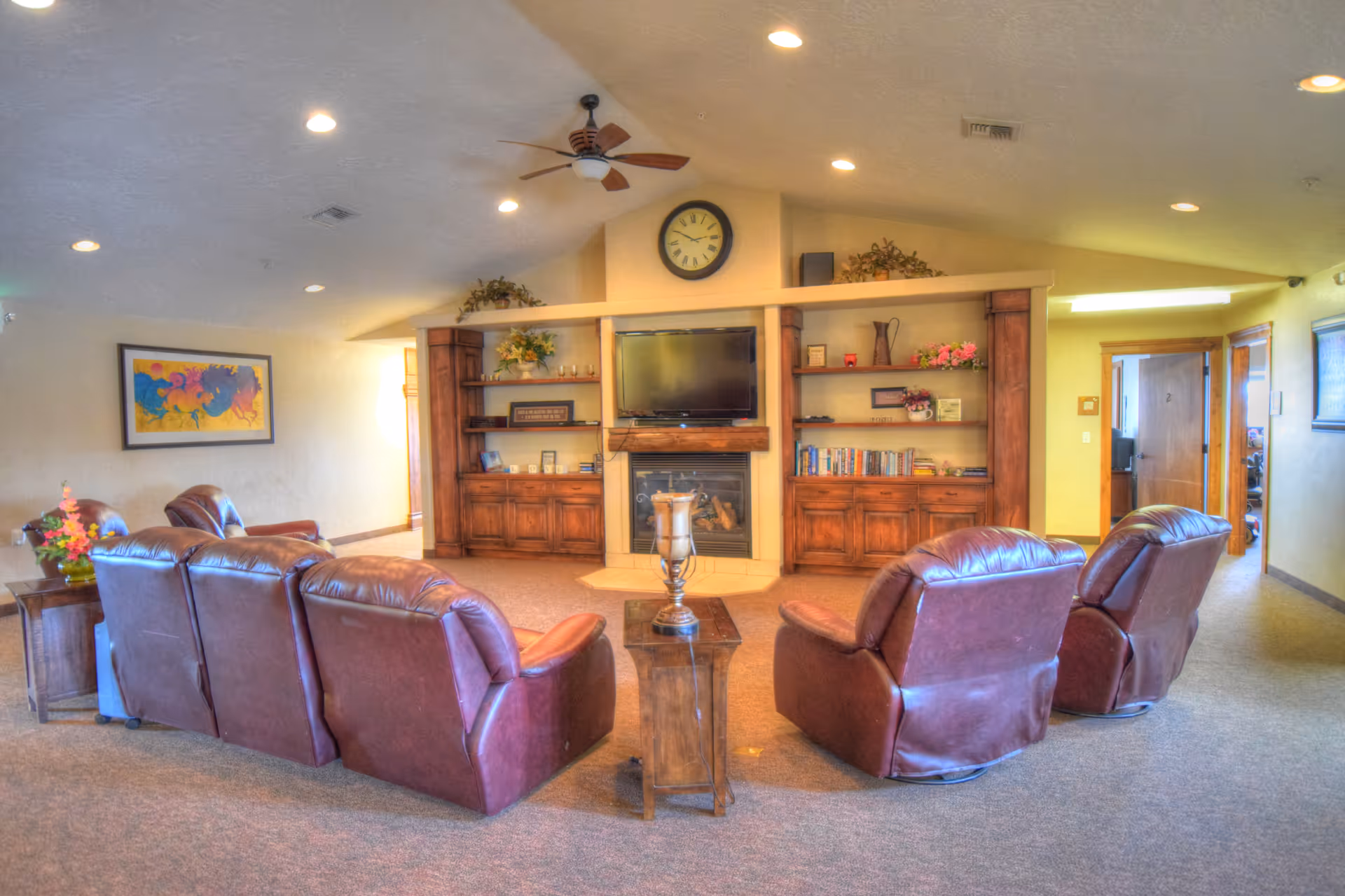 A cozy living room area with four brown leather recliners arranged in a semicircle facing a built-in wooden entertainment center with a flat-screen TV mounted above a fireplace. The entertainment center has shelves with books, decorative items, and plants. A ceiling fan with lights is mounted on the ceiling, and there is a colorful abstract painting on the left wall. The room is well-lit with recessed lighting and has carpeted flooring.
