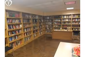 Interior view of a library room with wooden bookshelves filled with books lining two walls. There is a wooden desk with a chair in the corner and a table with some items on it in the foreground. The room has a tiled floor and a drop ceiling with fluorescent lighting.