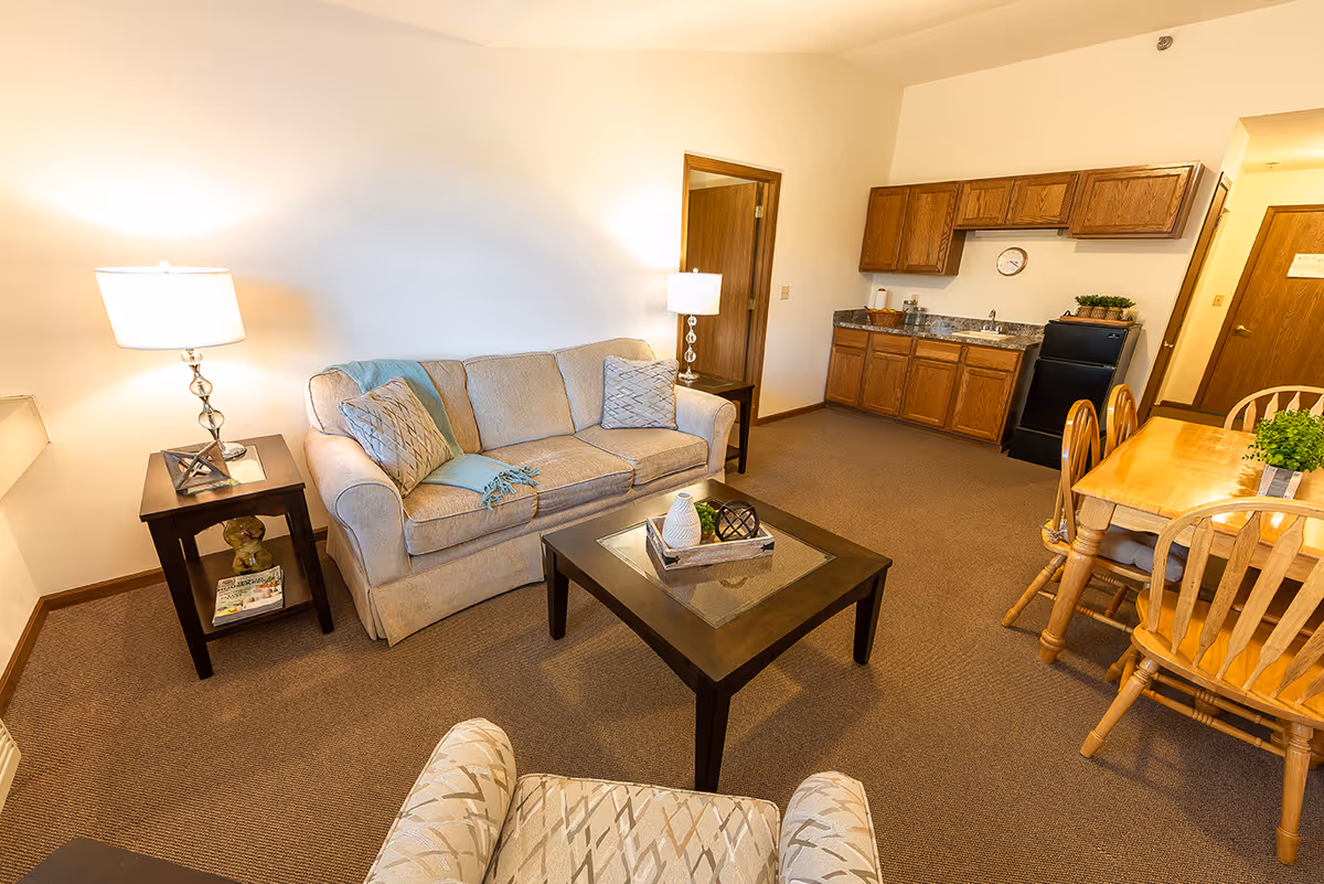 A cozy senior living room with a beige sofa, two side tables with lamps, a patterned armchair, a wooden coffee table with decorative items, a small kitchen area with wooden cabinets, a black mini fridge, and a wooden dining table with chairs. The room has beige carpet and white walls.