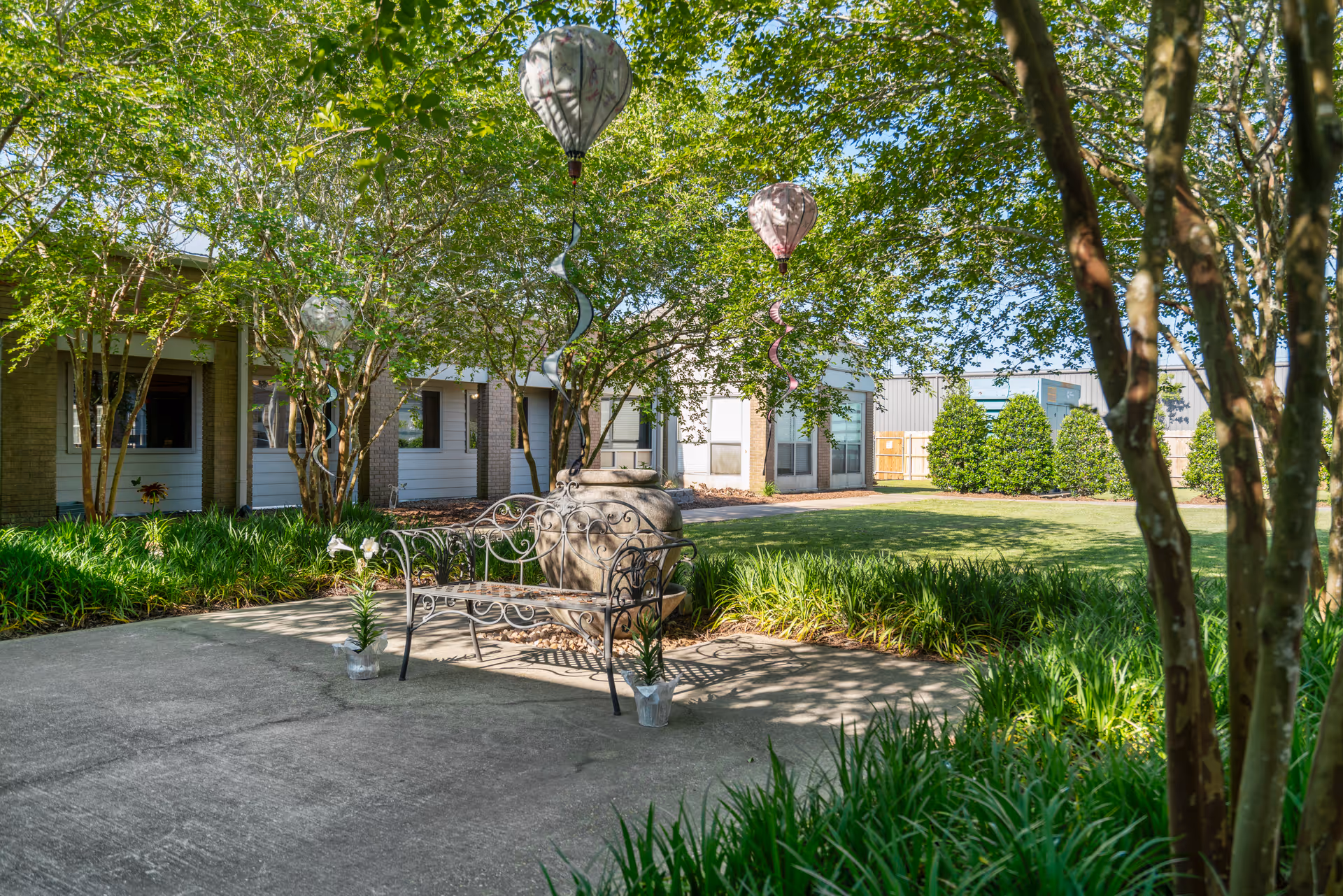 Outdoor garden area at Grand Cove featuring a metal bench on a concrete patio surrounded by green plants and trees. Decorative hanging ornaments shaped like hot air balloons are suspended from the tree branches. The background shows part of the building with windows and a well-maintained lawn.