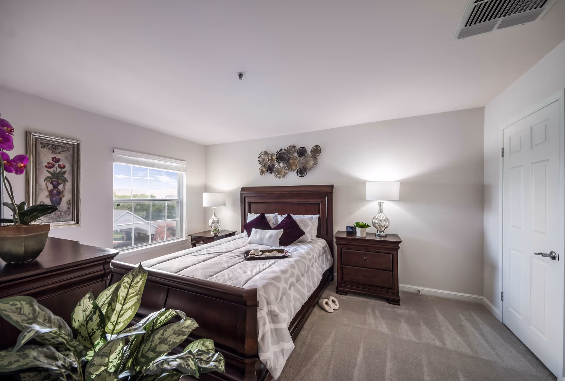 A bright bedroom with a large window letting in natural light. The room features a wooden bed with white and purple bedding, two matching wooden nightstands with lamps, a decorative wall piece above the bed, a framed floral painting on the wall, and several green plants adding a touch of nature to the space.