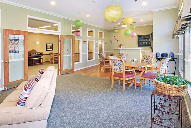 A bright and spacious common area in a senior living facility with a beige sofa adorned with striped pillows on the left, several wooden tables with floral cushioned chairs in the center, and colorful hanging paper lanterns from the ceiling. The room has large windows on the right side letting in natural light, and a small table with a basket of green plants near the window. In the background, there are glass-paneled double doors leading to another room with a piano and additional seating.