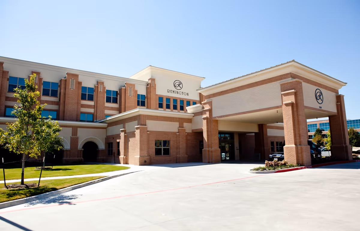 Exterior view of Remington Transitional Care of San Antonio, a multi-story brick building with large windows and a covered entrance driveway. The building is surrounded by a well-maintained lawn and small trees under a clear blue sky.