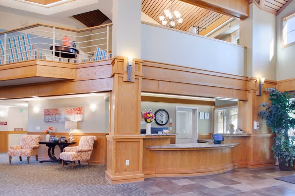 A spacious senior living facility lobby with a wooden reception desk, a large clock on the wall behind the desk, and a vase of flowers on the counter. There are two patterned armchairs with a small round table between them, decorated with flowers and picture frames. The area features wood paneling, a high ceiling with exposed beams, and a second-floor balcony with additional seating. A large potted plant is visible near the entrance.
