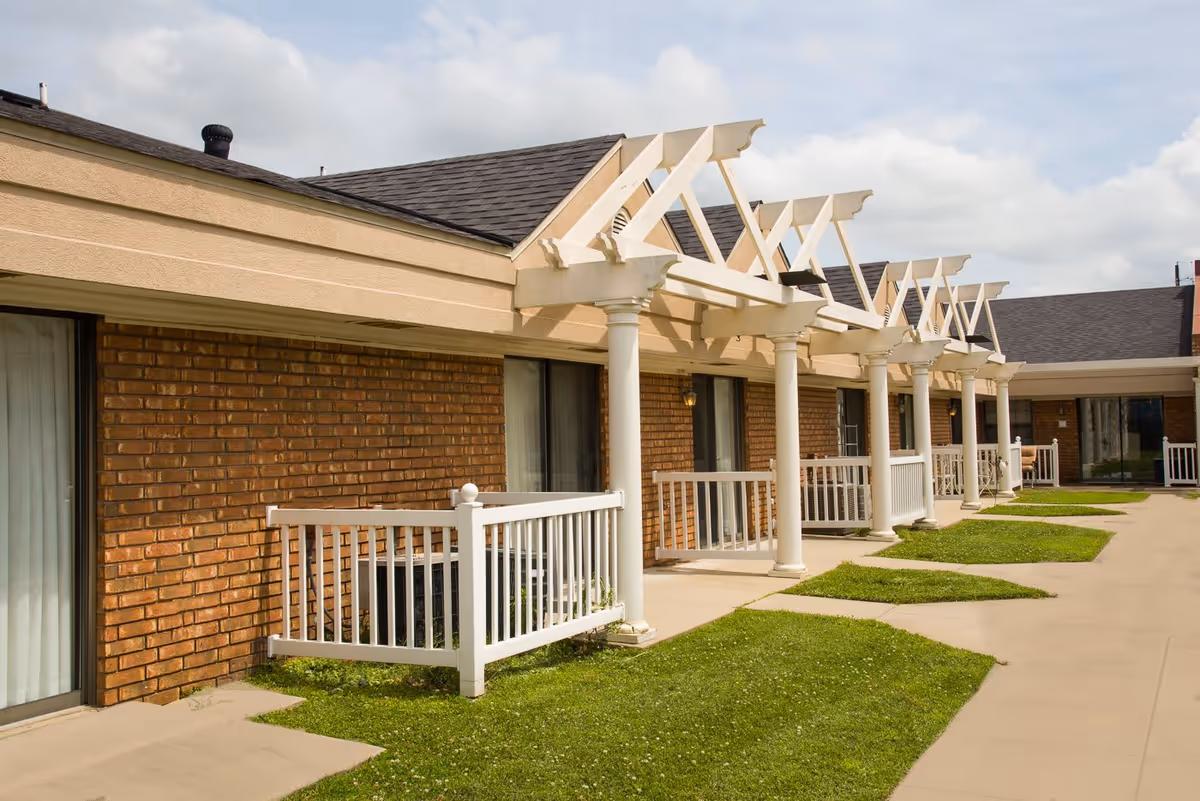Exterior view of a single-story assisted living facility with brick walls, white columns, and small fenced patios in front of sliding glass doors. The building has a dark shingled roof and a paved walkway with patches of green grass.