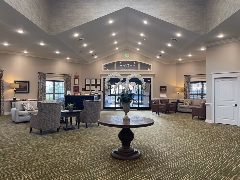 Spacious and well-lit senior living facility lobby with a round wooden table holding a vase of white orchids in the center. The room features comfortable seating areas with armchairs and sofas on either side, large windows with curtains, framed artwork on the walls, and a reception desk in the background near the entrance doors.