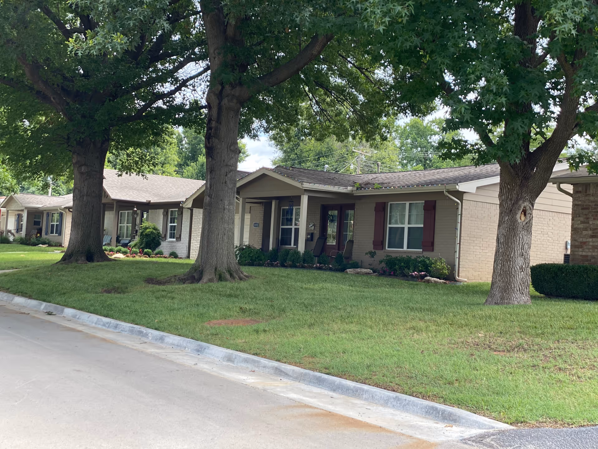 Exterior view of single-story residential buildings with large trees and well-maintained lawns in front, under a partly cloudy sky.