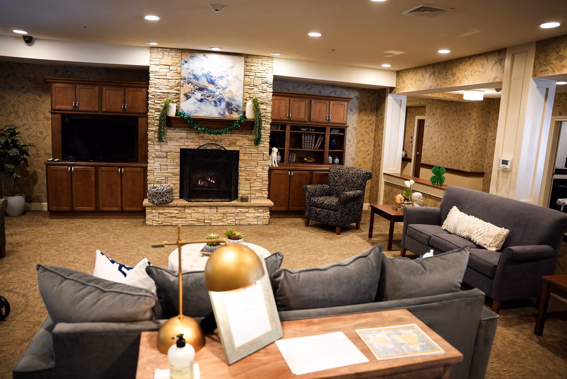 A cozy living room area in a senior living facility featuring a stone fireplace with a painting above it and green garland decorations. The room has wooden cabinets on either side of the fireplace, a patterned armchair, a gray sofa with a white pillow, a wooden side table with flowers, and a desk with a gold lamp and hand sanitizer in the foreground. The walls have a floral wallpaper and the ceiling has recessed lighting.
