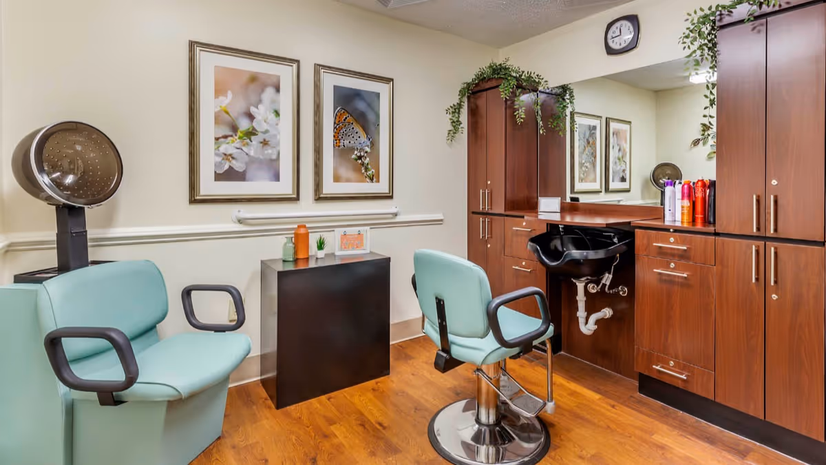 Interior of a salon area in a senior living facility featuring two light green salon chairs, a hair washing station with a black sink, wooden cabinets, a hair dryer, and two framed pictures of flowers and a butterfly on the wall.