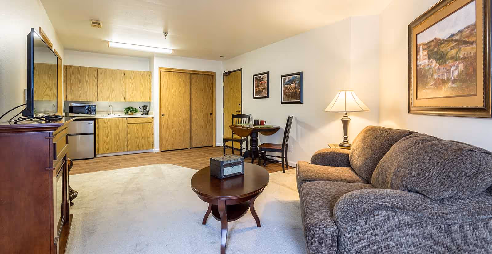 A cozy living area in a senior living facility featuring a gray upholstered sofa, a wooden coffee table with a decorative box, a small round dining table with two chairs, and a kitchenette with wooden cabinets, a microwave, and a mini fridge. The room is softly lit by a table lamp and has framed artwork on the walls.
