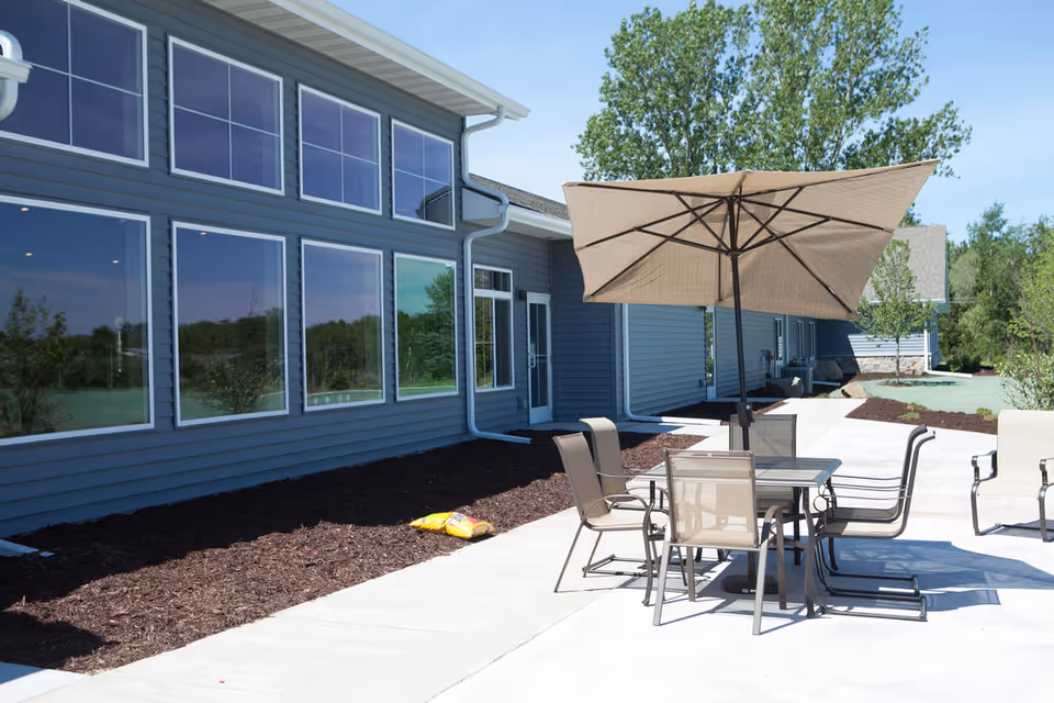 Patio area with a table, chairs and a large umbrella next to a modern building's row of large windows.